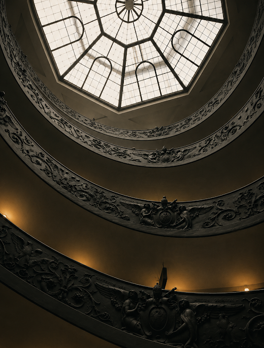 Spiral staircase with decorative elements and a large stained glass window at the Vatican Museum.